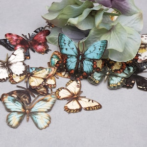May include: A collection of decorative butterfly ornaments in various colors, including white, blue, red, and yellow, are arranged on a gray surface. Some butterflies are near green and purple floral accents. The butterflies have detailed wing patterns.