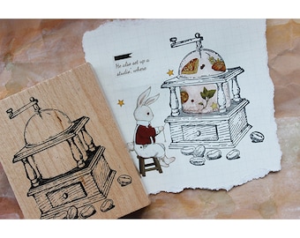 May include: A wooden stamp with a black and white illustration of a coffee grinder. The stamp is on a white background with a drawing of a coffee grinder, a rabbit, and coffee beans. The text "He also set up a studio, where" is written on the background.