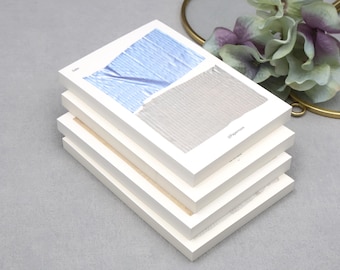 May include: A stack of white notebooks with abstract art on the top cover. The artwork features blue and grey brushstrokes. The notebooks are stacked on a grey surface, with a decorative gold ring and flowers in the background.