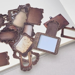 May include: A collection of vintage-style brown paper frames and labels with ornate, decorative edges. Some frames have blank spaces, while others have a distressed, aged appearance. The frames are scattered on a white surface.