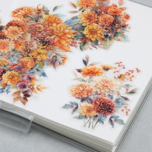 May include: A notebook with floral stickers featuring orange, yellow, and pink dahlias and other flowers. The stickers are arranged in clusters and have a watercolor effect. The notebook is white with a clear plastic cover.