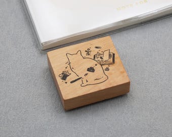 May include: A wooden stamp with a cat illustration. The stamp features a cat outline with a book, coffee cup, and other small objects. The stamp is on a gray surface with a white notebook in the background. The stamp is square.