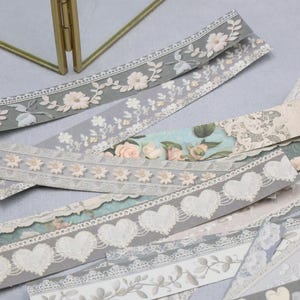 Op de afbeelding: Assortiment van decoratieve papieren linten met bloemen-, hart- en kanten ontwerpen. De linten hebben verschillende kleuren, waaronder grijs, wit en pasteltinten. Sommige linten hebben reliëf bloempatronen, andere delicate kanten randen.