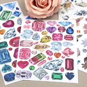 May include: Sheets of colorful gem stickers in various shapes and sizes, including emerald cuts, hearts, and ovals. The gems are in shades of blue, pink, green, and yellow. A pink rose is in the background.
