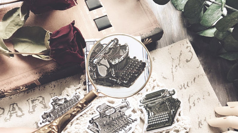 May include: A close-up of a vintage typewriter sticker with a magnifying glass over it. The sticker is on a white background with other vintage typewriter stickers.