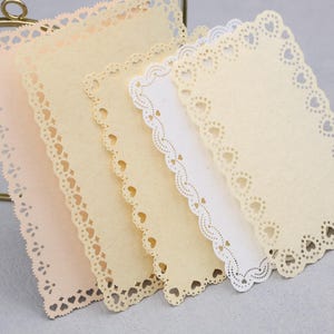Könnte beinhalten: Ein Set aus fünf rechteckigen Papierkarten mit gestanzten herzförmigen Rändern. Die Karten haben verschiedene Nuancen von Creme, Beige und Weiß, jede mit einem zarten Wellenrand und einem wiederkehrenden Muster aus kleinen Herzstanzungen.