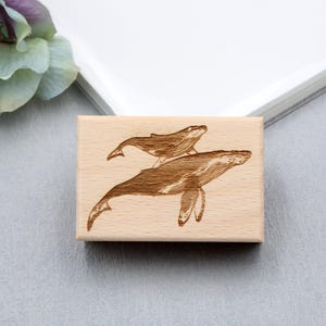 May include: A rectangular wooden stamp featuring an engraved design of two whales. The larger whale is positioned below, with a smaller whale above it. The whales are depicted in a detailed, artistic style, with visible lines and shading.