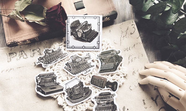 May include: A set of vintage typewriter stickers in black and white. The stickers are on a white background with a lace doily underneath. The stickers are perfect for adding a vintage touch to your journals, planners, or scrapbooks.