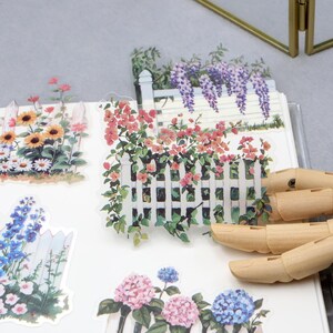 May include: A collection of floral stickers featuring white picket fences and various flowers, including sunflowers, bluebells, and hydrangeas. The stickers are arranged on a white surface, with a wooden hand in the foreground.