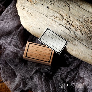 May include: Two wooden stamps with black frames and dotted lines. The stamps are on a grey fabric background. The larger stamp is 50 x 30 mm.