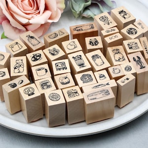 May include: A collection of wooden stamps with various food-related illustrations, including a bear, coffee cup, and fish. The stamps are arranged on a white plate, with a pink rose in the background. The stamps are made of light-colored wood.