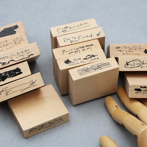 May include: A collection of rectangular wooden rubber stamps with various handwritten phrases and ink markings on the sides. The stamps feature text such as Coffee and Chill, Daily Coaster, and other illegible cursive scripts, arranged in a cluster.