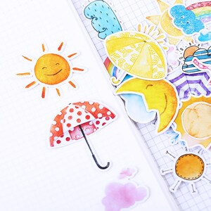 May include: A sheet of colorful stickers featuring various weather-themed illustrations, including a smiling sun, a red umbrella with white polka dots, a yellow umbrella with green leaves, a crescent moon, and a cloud with a rainbow.