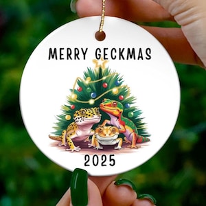 Peut inclure: Ornement de Noël rond et blanc avec l'inscription "MERRY GECKMAS" et l'année "2025". L'ornement représente un sapin de Noël et trois geckos jaunes, rouges et verts. L'ornement est tenu par une main.