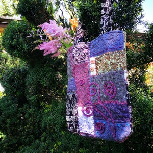 Può includere: Una borsa a tracolla in patchwork realizzata con diversi tessuti in tonalità di viola, rosa, marrone e nero. La borsa ha una chiusura a bottone e un ricamo floreale sul davanti.