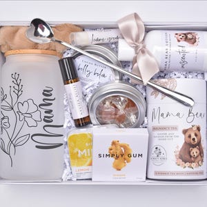 May include: A white gift box filled with self-care items. Includes a frosted glass jar with the word "Mama", a silver spoon, lotion, tea, and ginger gum. The box is decorated with a ribbon and floral accents.