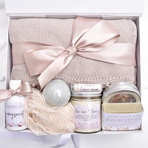 Op de afbeelding: Een geschenkdoos met een beige gebreide deken vastgebonden met een satijnen lint, een fles hand- en bodylotion, een net-spons, een ronde badbom, een citroen lippenbalsem, een kaars met tekst en een stuk eucalyptus shea zeep in een verpakking.