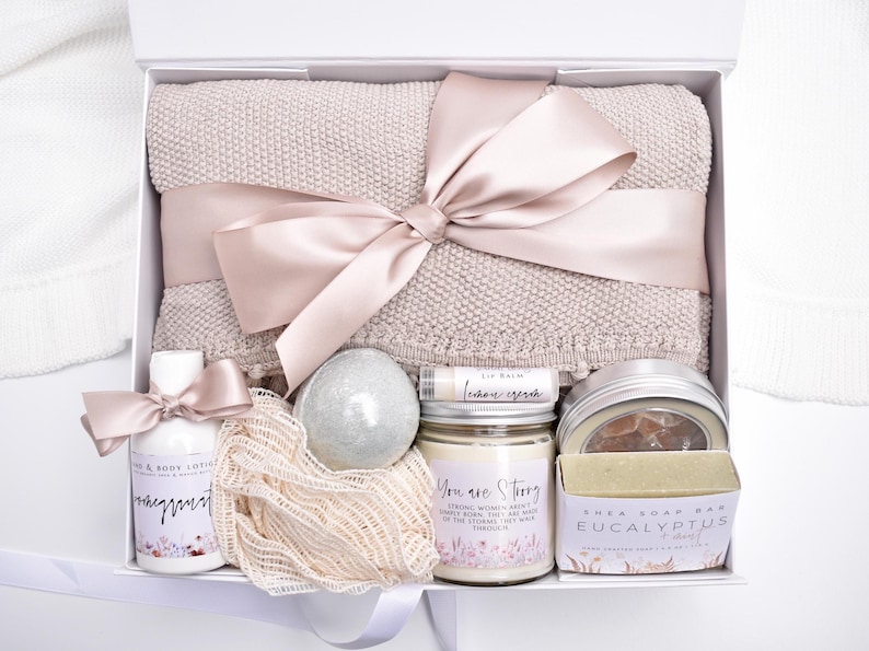 Op de afbeelding: Een geschenkdoos met een beige gebreide deken vastgebonden met een satijnen lint, een fles hand- en bodylotion, een net-spons, een ronde badbom, een citroen lippenbalsem, een kaars met tekst en een stuk eucalyptus shea zeep in een verpakking.