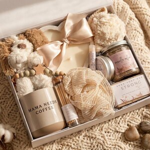 May include: A gift box containing a plush puppy, a heart-shaped candle with a satin ribbon, a mug labeled MAMA NEEDS COFFEE, a jar of vanilla coffee scrub, a shea soap bar labeled STRONG, a mesh bath sponge, a lip balm, and a small bottle of matches.