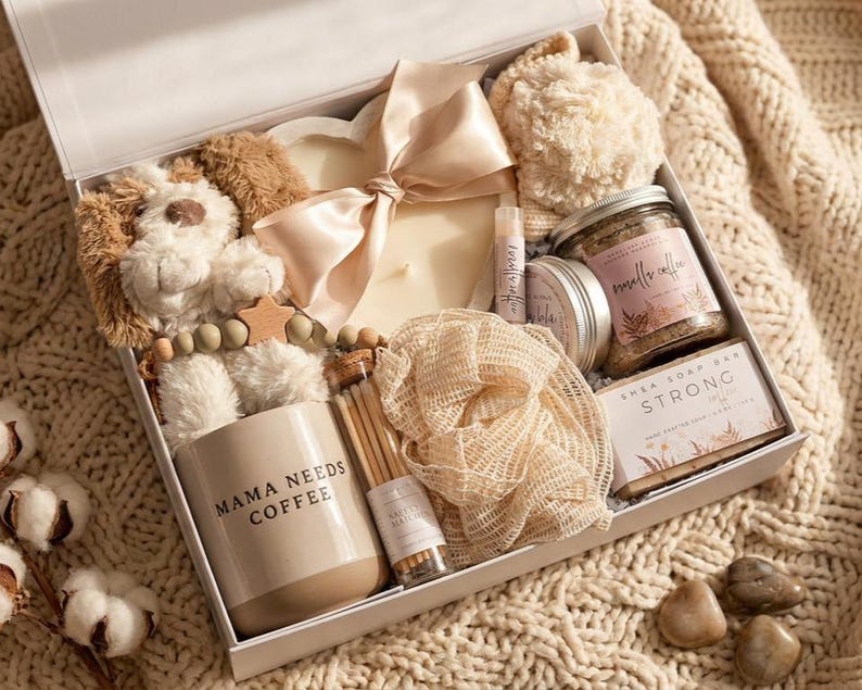 May include: A gift box containing a plush puppy, a heart-shaped candle with a satin ribbon, a mug labeled MAMA NEEDS COFFEE, a jar of vanilla coffee scrub, a shea soap bar labeled STRONG, a mesh bath sponge, a lip balm, and a small bottle of matches.