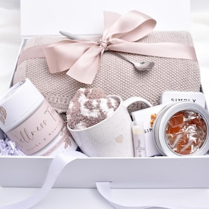 May include: A white gift box filled with self-care items. Includes a blanket, mug with socks, a jar labeled "Wellness Tea", lip balm, a tin of orange-colored items, and a small card. The box is tied with a pink ribbon.