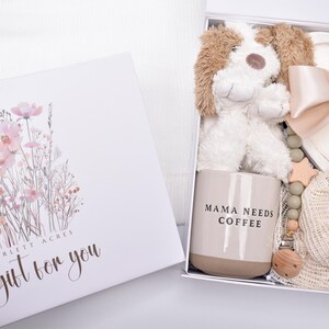 May include: A white gift box with a floral illustration and the text Scarlett Acres A gift for you. Inside is a plush puppy, a beige mug with the text MAMA NEEDS COFFEE, a heart-shaped candle with a satin ribbon, and a wooden bead pacifier clip.