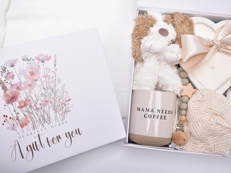 May include: A white gift box with a floral illustration and the text Scarlett Acres A gift for you. Inside is a plush puppy, a beige mug with the text MAMA NEEDS COFFEE, a heart-shaped candle with a satin ribbon, and a wooden bead pacifier clip.
