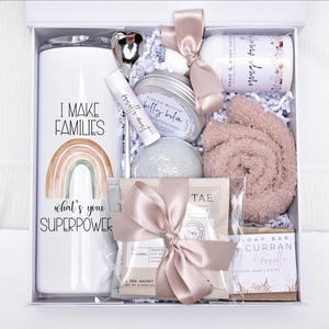 May include: A white gift box filled with self-care items. Includes a tumbler with a rainbow graphic and the text "I MAKE FAMILIES, what's your SUPERPOWER", a lip balm, a heart-shaped spoon, a candle, a soap bar, a pair of socks, and a tea sachet.