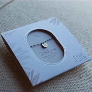 May include: A sterling silver charm bracelet on a thin dark cord, presented in a square white box with a central oval cutout. The packaging features the text "EVER ASTER", "STERLING SILVER", and "thank you FOR ALL THAT YOU DO" printed in black.