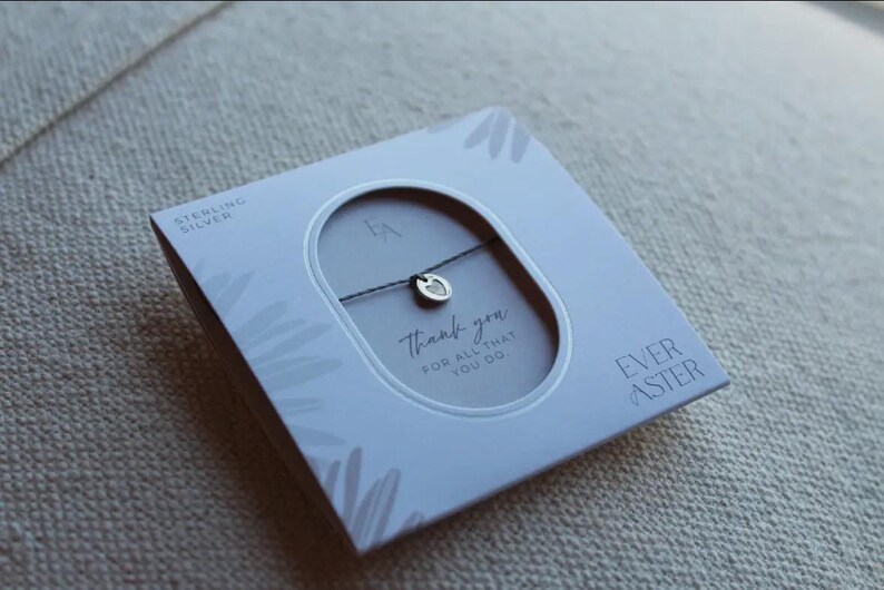 May include: A sterling silver charm bracelet on a thin dark cord, presented in a square white box with a central oval cutout. The packaging features the text "EVER ASTER", "STERLING SILVER", and "thank you FOR ALL THAT YOU DO" printed in black.