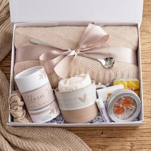 May include: A gift box containing a beige knit blanket tied with a satin ribbon and a silver spoon, a Wellness Tea canister, a ceramic mug with a heart, a white scrunchie, a lip balm, and a round tin with orange candies inside a clear window.