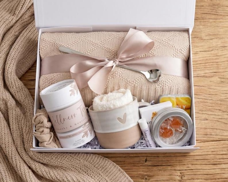 May include: A gift box containing a beige knit blanket tied with a satin ribbon and a silver spoon, a Wellness Tea canister, a ceramic mug with a heart, a white scrunchie, a lip balm, and a round tin with orange candies inside a clear window.