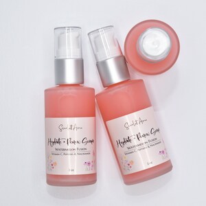 Op de afbeelding: Twee flessen van 2 ounce roze hydraterende en vernieuwende serum met een watermeloenfusie, vitamine C, peptiden en niacinamide. De flessen hebben een zilveren pomptop en een wit etiket met roze bloemmotieven.