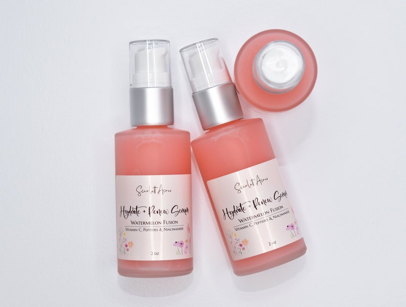 Op de afbeelding: Twee flessen van 2 ounce roze hydraterende en vernieuwende serum met een watermeloenfusie, vitamine C, peptiden en niacinamide. De flessen hebben een zilveren pomptop en een wit etiket met roze bloemmotieven.