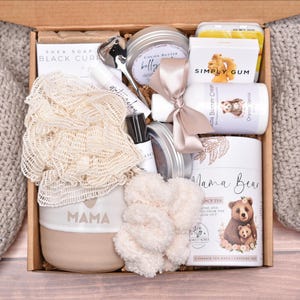 May include: A gift box containing a beige ceramic mug with the word MAMA, a cream-colored mesh bath sponge, a white Simply Gum box, a Mama Bear tea canister, a cocoa butter cream bottle with a beige ribbon, a metal spoon, and various skincare items.