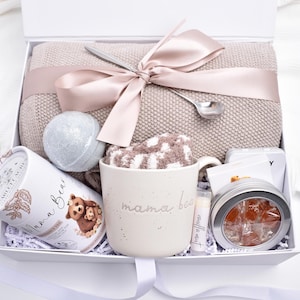 May include: A gift box filled with self-care items. Includes a beige blanket tied with a pink ribbon, a mug with "mama bear" text, a bath bomb, socks, tea, lip balm, and a tin of candies. The box is white with a white ribbon.