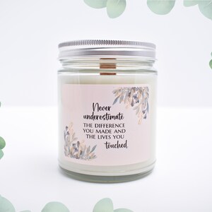 May include: A clear glass candle jar with a silver metal lid and a wooden wick. The label features a light pink background with blue and gold floral illustrations and the text: Never underestimate THE DIFFERENCE YOU MADE AND THE LIVES YOU touched.