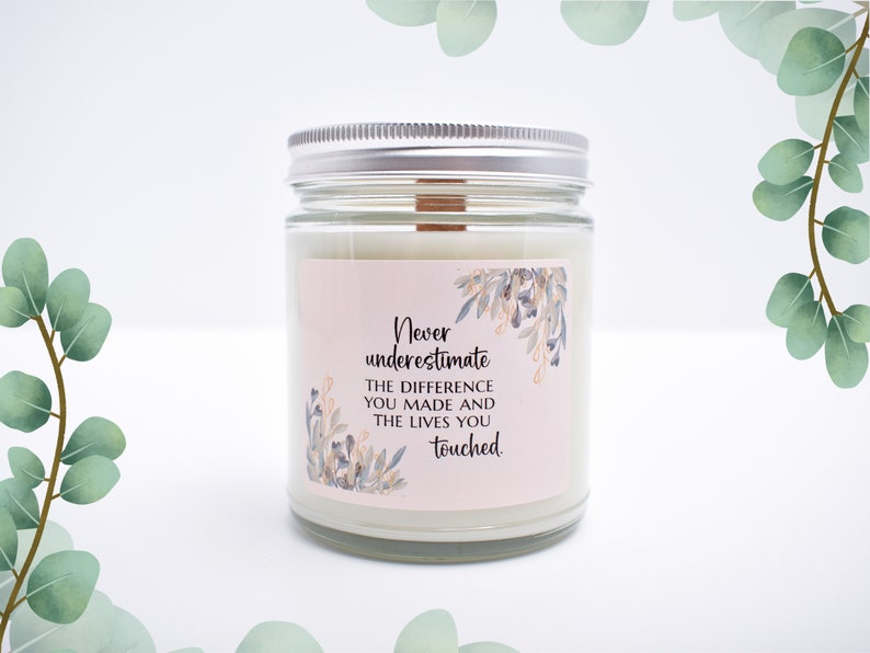 May include: A clear glass candle jar with a silver metal lid and a wooden wick. The label features a light pink background with blue and gold floral illustrations and the text: Never underestimate THE DIFFERENCE YOU MADE AND THE LIVES YOU touched.