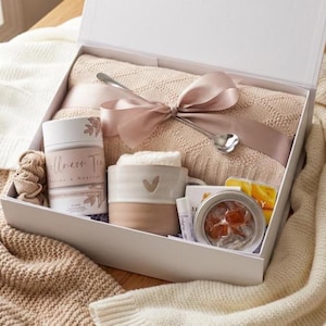May include: A white gift box containing a folded beige knit blanket tied with a satin ribbon, a silver spoon, a cylindrical container labeled Wellness Tea, a two-toned ceramic mug with a heart detail, a lip balm, and a small tin of candies.
