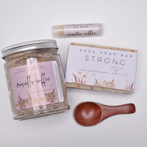 May include: A set containing a glass jar of whipped sugar scrub labeled Vanilla Coffee, a rectangular shea soap bar labeled Strong Coffee, a cylindrical lip balm labeled Vanilla Coffee, and a small wooden scoop with a dark brown finish.