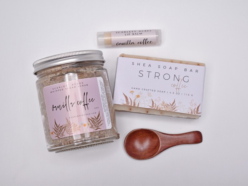 May include: A set containing a glass jar of whipped sugar scrub labeled Vanilla Coffee, a rectangular shea soap bar labeled Strong Coffee, a cylindrical lip balm labeled Vanilla Coffee, and a small wooden scoop with a dark brown finish.