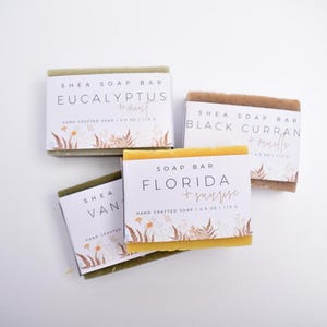 Op de afbeelding: Vier rechthoekige shea zeepblokken met witte papieren labels voorzien van bloemillustraties. De labels bevatten de tekst Eucalyptus + Mint, Black Currant + Vanilla, Florida + Sunrise en Vanilla, elk met de vermelding van 113 g.