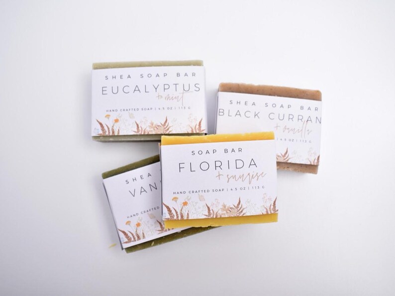 Op de afbeelding: Vier rechthoekige shea zeepblokken met witte papieren labels voorzien van bloemillustraties. De labels bevatten de tekst Eucalyptus + Mint, Black Currant + Vanilla, Florida + Sunrise en Vanilla, elk met de vermelding van 113 g.