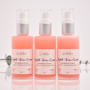 Op de afbeelding: Drie flessen roze serum van 2 ounce met het label "Hydrate + Renew Serum" met een bloemenmotief en de tekst "Watermelon Fusion, Vitamin C, Peptides & Niacinamide".
