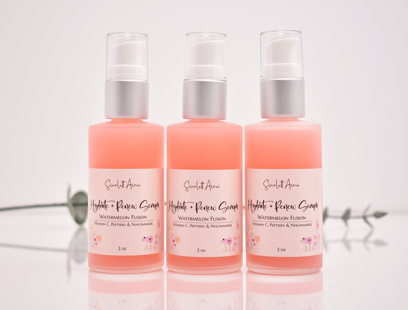 Op de afbeelding: Drie flessen roze serum van 2 ounce met het label "Hydrate + Renew Serum" met een bloemenmotief en de tekst "Watermelon Fusion, Vitamin C, Peptides & Niacinamide".