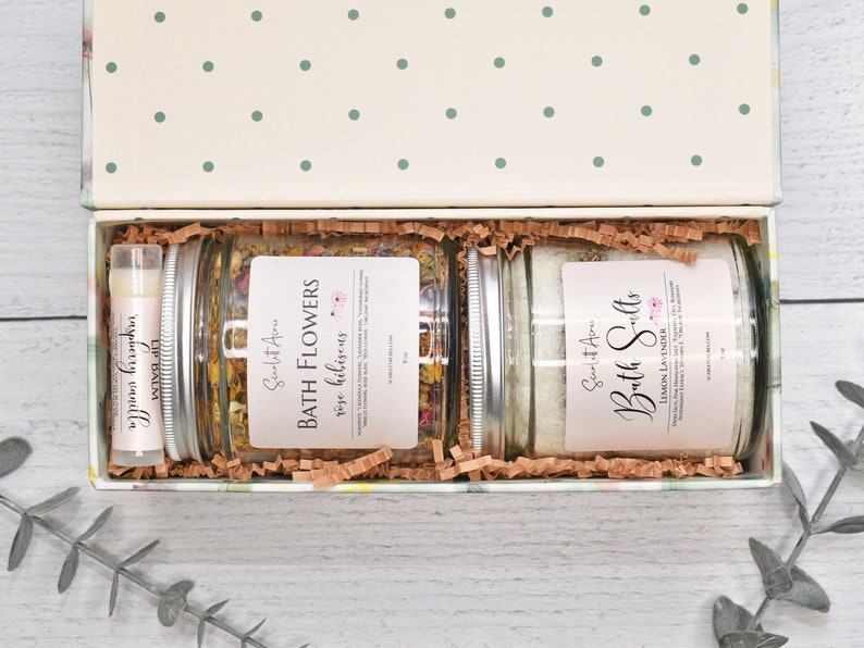 May include: A gift box filled with bath products. The box is open and contains two glass jars with labels that read "Scent It All Bath Flowers rose hibiscus" and "Scent It All Bath Salts Lemon Lavender". There is also a small tube of lip balm.