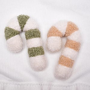 May include: Two candy cane shaped ornaments made of textured sherpa fabric. One ornament features alternating stripes of cream white and sage green, while the other features alternating stripes of cream white and light tan beige.
