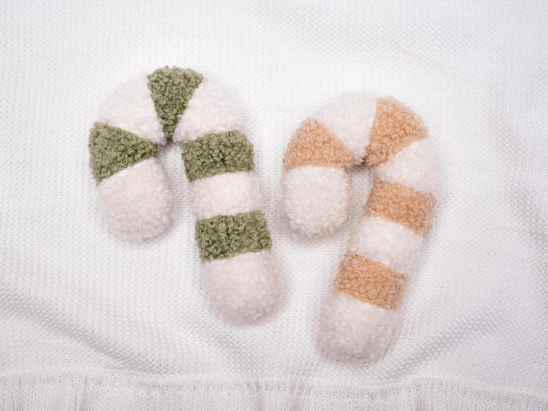 May include: Two candy cane shaped ornaments made of textured sherpa fabric. One ornament features alternating stripes of cream white and sage green, while the other features alternating stripes of cream white and light tan beige.