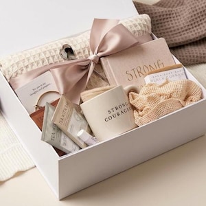 May include: A white gift box containing a beige waffle-knit blanket tied with a mauve ribbon, a notebook with the word STRONG, a mug labeled STRONG & COURAGEOUS, a black currant soap bar, tea packets, a lip balm, and a beige mesh scrunchie.