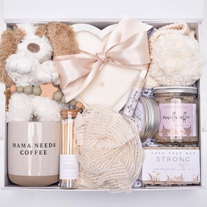 May include: A gift box containing a plush dog toy, a heart-shaped candle with a satin ribbon, a mug labeled MAMA NEEDS COFFEE, a glass tube of matches, a mesh sponge, a lip balm, a jar of vanilla coffee scrub, and a bar of shea soap labeled STRONG COFFEE.